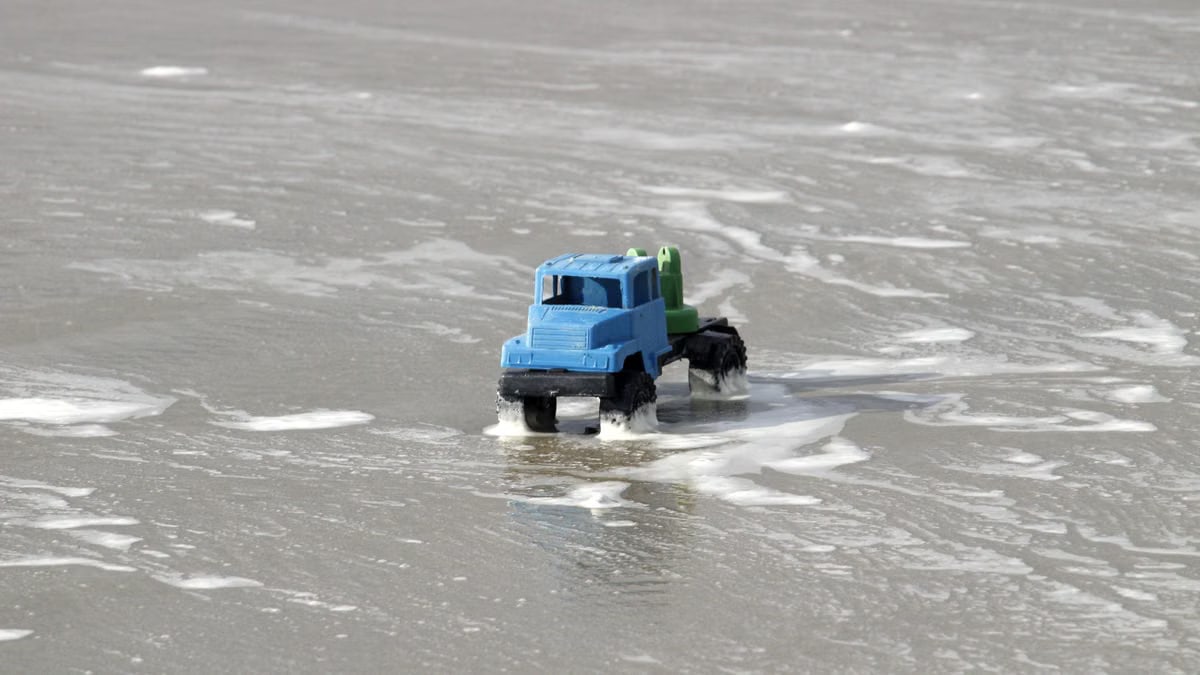 A blue toy truck navigates foamy ocean waves on Guaratuba Beach, Brazil.